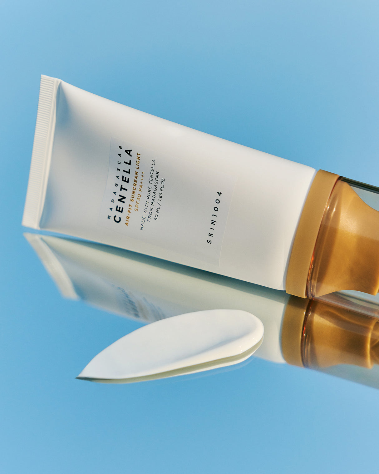 Il tubetto bianco con tappo dorato trasparente del “Skin1004 Madagascar Centella Air-Fit Suncream Light” è disteso su una superficie riflettente azzurra, accanto a una generosa quantità di crema bianca applicata a spatola.