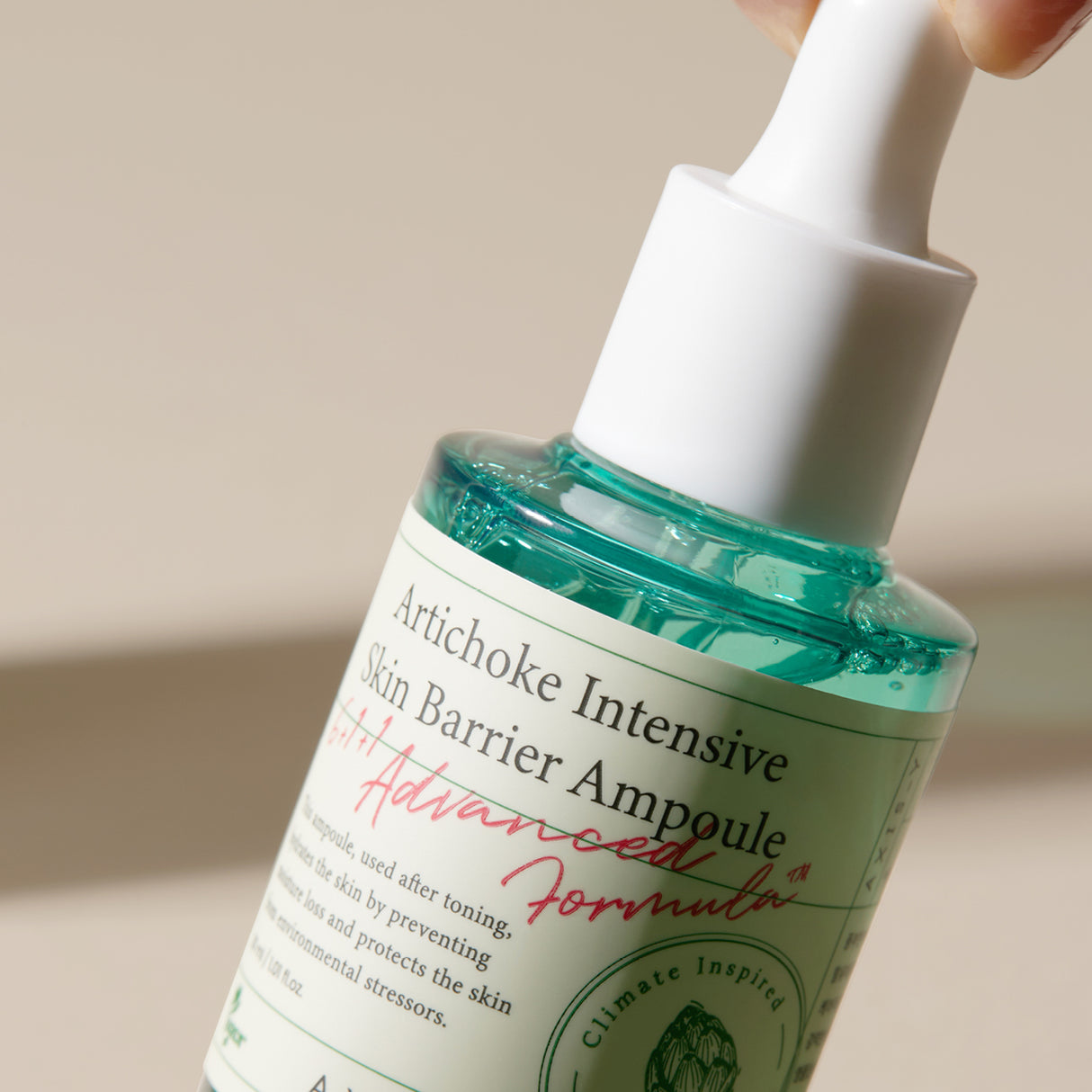 Primo piano inclinato del flacone in vetro del AXIS-Y Artichoke Intensive Skin Barrier Ampoule, con liquido azzurro e tappo contagocce bianco. L’etichetta mostra chiaramente il nome del prodotto e la scritta “Advanced Formula” in rosso. Sfondo neutro beige.