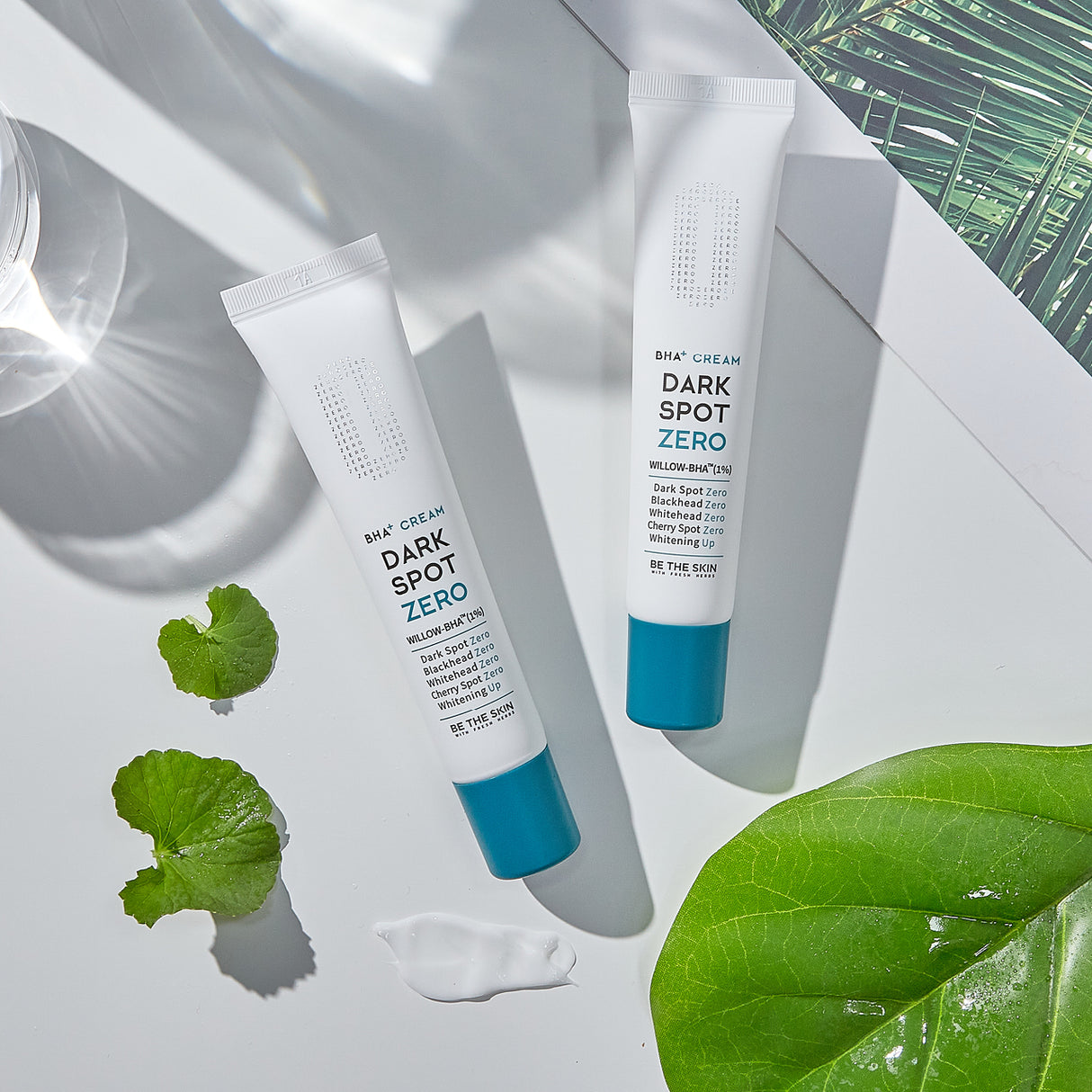 Due tubi di crema BE THE SKIN Dark Spot Zero BHA+ su sfondo bianco, accompagnati da foglie di centella asiatica, una foglia verde brillante e schizzi di crema.