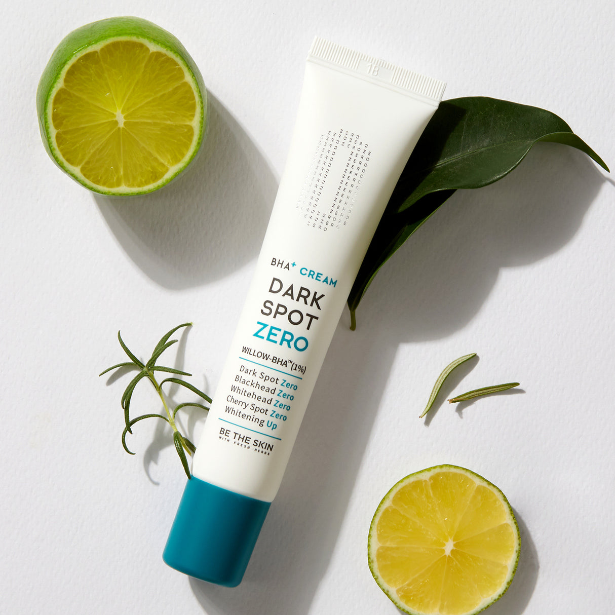 Tubo di crema BE THE SKIN Dark Spot Zero BHA+ su sfondo bianco, circondato da foglie verdi, rosmarino e fette di lime, con il tappo azzurro in evidenza.