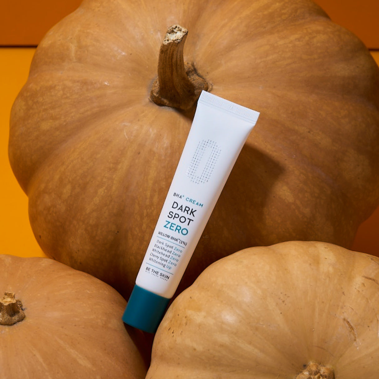Tubo della crema BE THE SKIN Dark Spot Zero BHA+ con tappo azzurro appoggiato su grandi zucche arancioni, in un setting autunnale dal tono caldo.