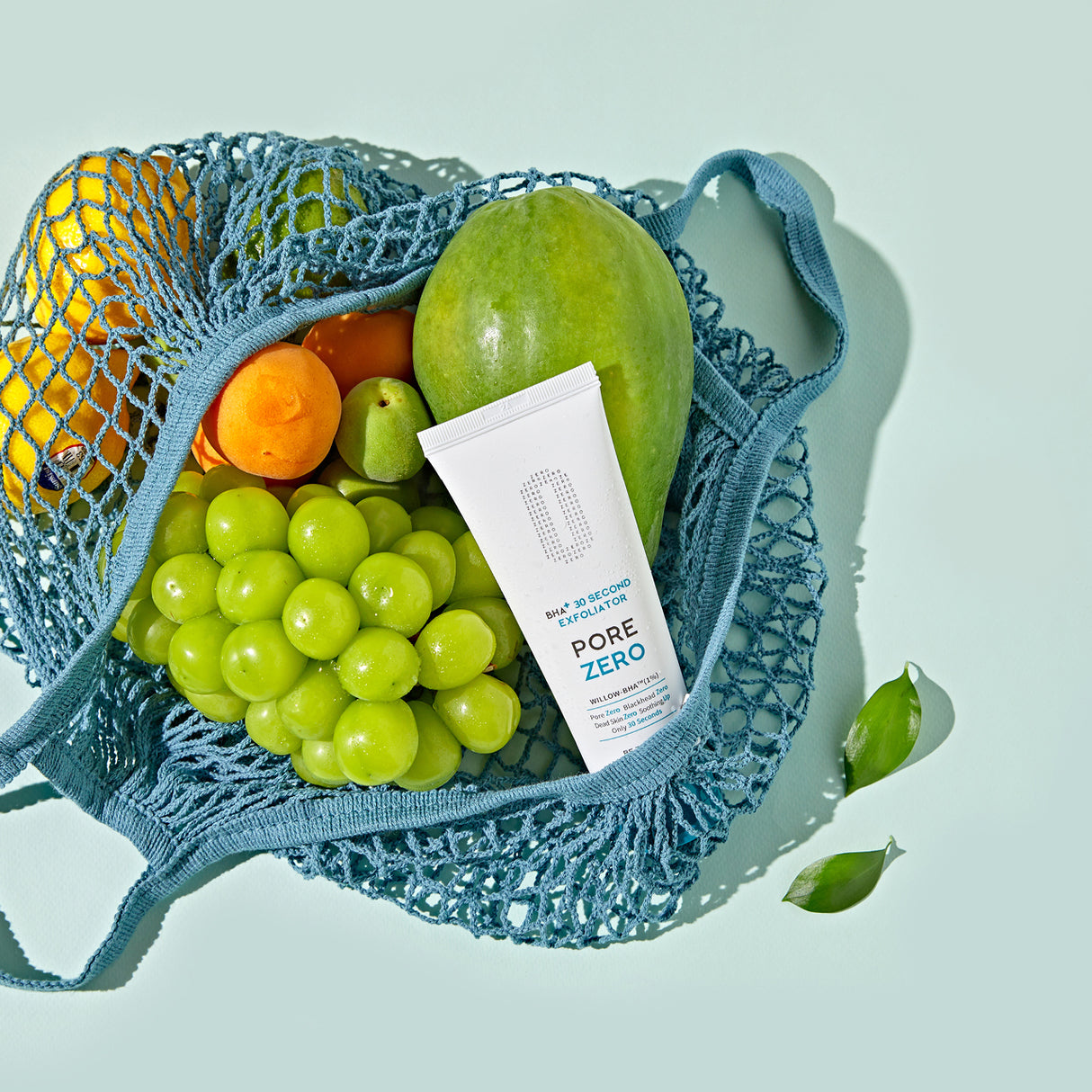 Tubo dell’esfoliante BE THE SKIN PORE ZERO BHA+ 30 Second Exfoliator inserito in una rete blu tra frutta fresca come uva, mango, albicocche e limone, su sfondo verde menta.