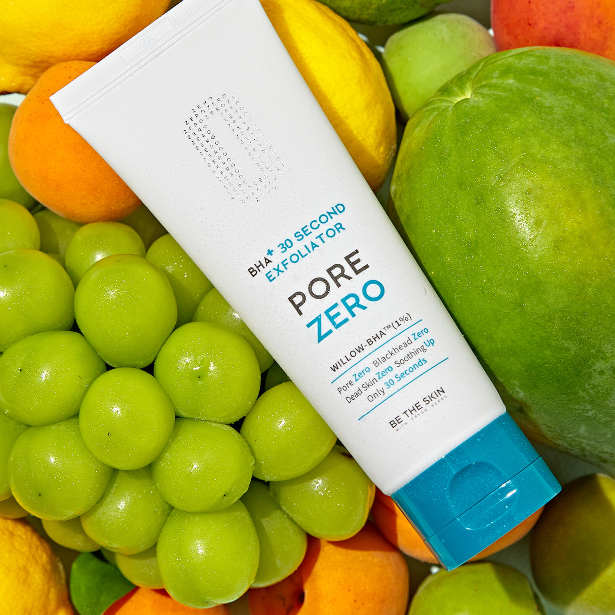 Tubo dell’esfoliante BE THE SKIN PORE ZERO BHA+ 30 Second Exfoliator appoggiato su un assortimento di frutta fresca, tra cui uva verde, mango, albicocche e agrumi, in un’ambientazione luminosa e vivace.