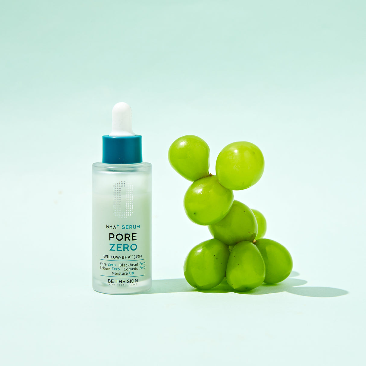 Flacone del siero BE THE SKIN PORE ZERO BHA+ con contagocce, posizionato accanto a un grappolo di uva verde su sfondo verde pastello, evocando freschezza e ingredienti naturali.