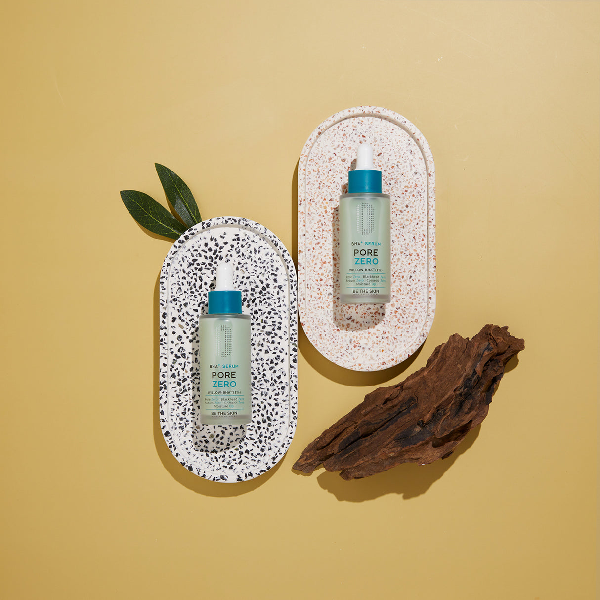 Due flaconi del siero BE THE SKIN PORE ZERO BHA+ disposti su piattini ovali con effetto terrazzo, su sfondo giallo senape, decorati con foglie verdi e un pezzo di corteccia.
