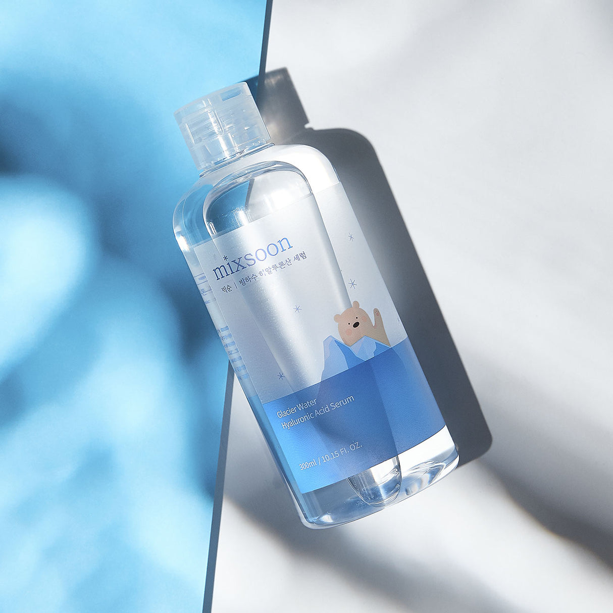 Flacone trasparente del siero Mixsoon Glacier Water Hyaluronic Acid Serum da 300 ml, adagiato su una superficie riflettente con giochi di luce azzurra e ombra, decorato con un'illustrazione di un orsetto tra i ghiacci.