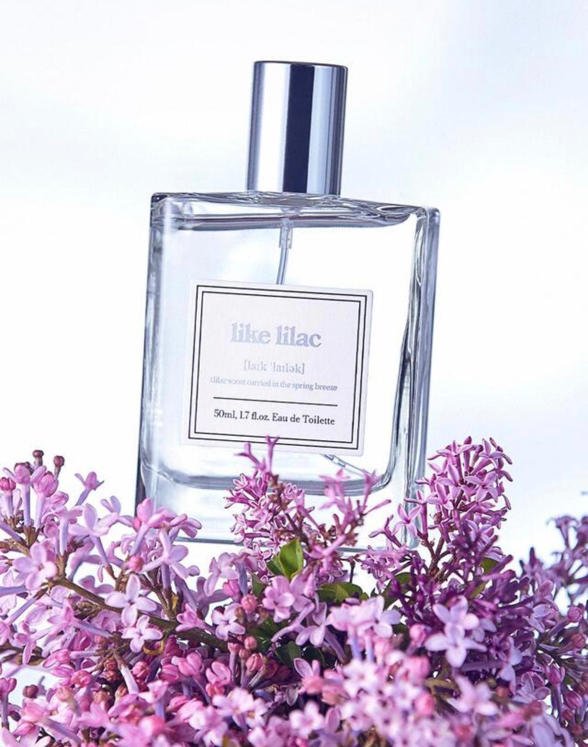 Flacone di profumo Like Lilac Eau de Toilette 50 ml in vetro trasparente con tappo argentato, fotografato su sfondo chiaro con fiori di lillà viola in primo piano.