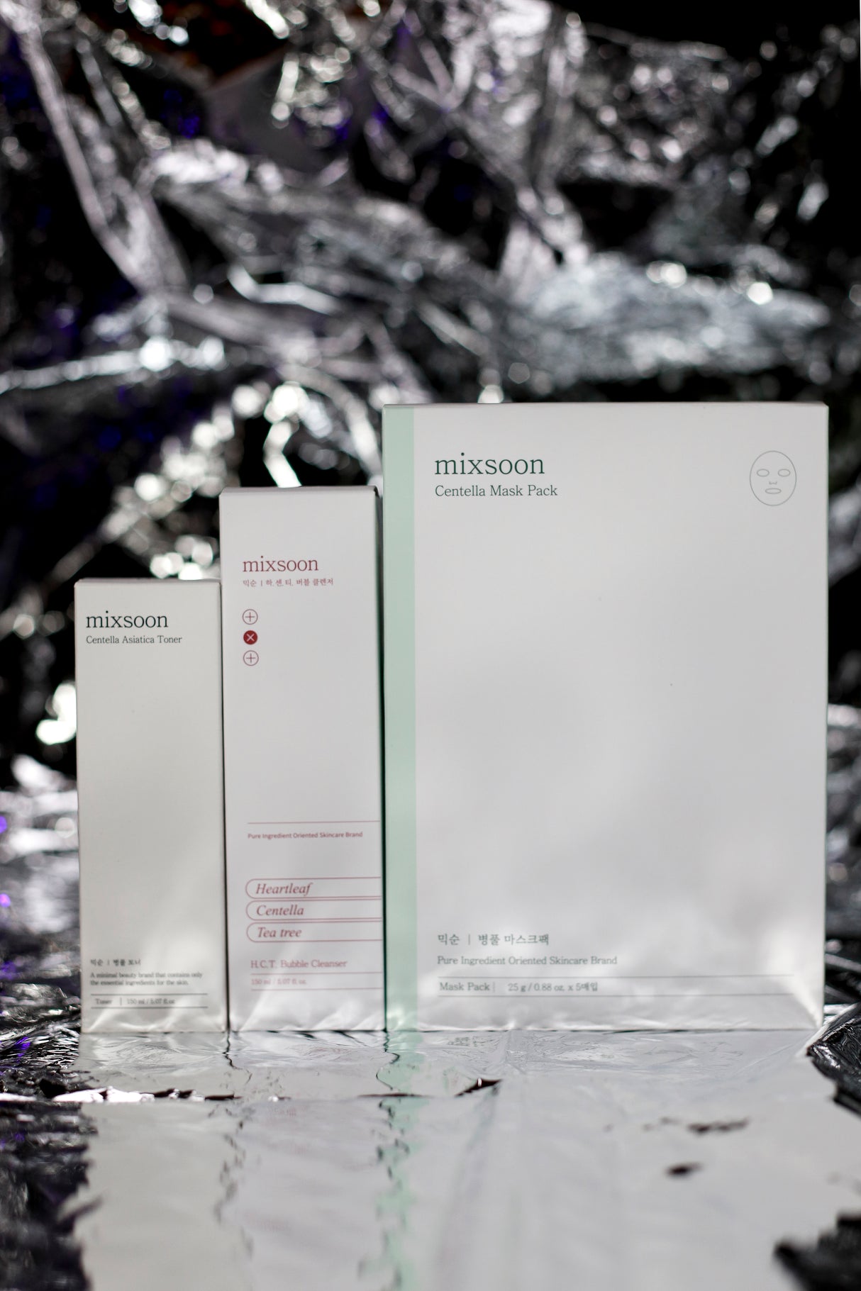 "Linea skincare 'mixsoon' con prodotti a base di ingredienti puri: tonico alla Centella Asiatica, detergente schiumogeno H.C.T. con Heartleaf, Centella e Tea Tree, e confezione di maschere Centella Mask Pack, presentati su sfondo argentato riflettente."