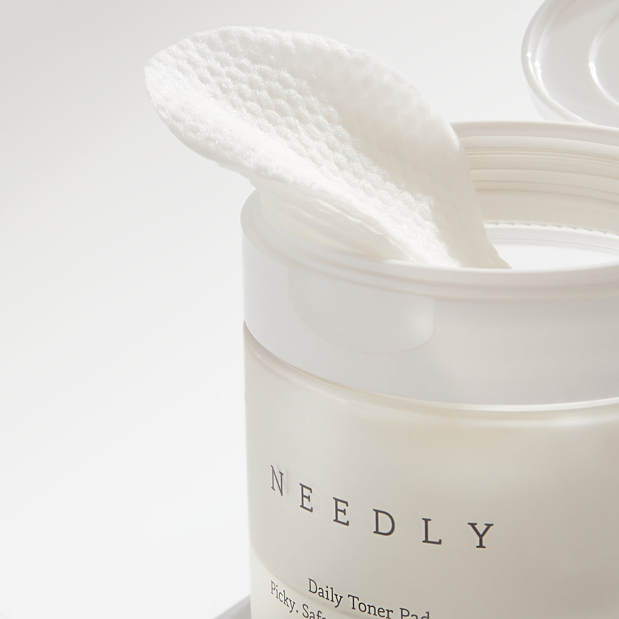 Barattolo aperto del "Needly Daily Toner Pad" con un dischetto esfoliante bianco dalla texture a nido d’ape parzialmente estratto, evidenziato da una luce naturale che ne mette in risalto la consistenza.