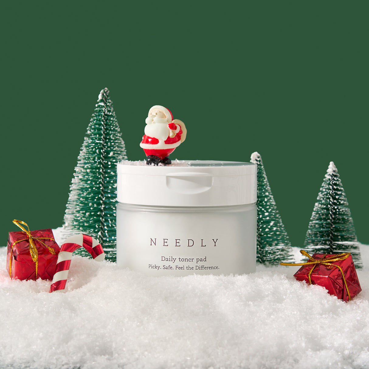 Vasetto di NEEDLY Daily Toner Pad posizionato su neve artificiale, circondato da decorazioni natalizie come pacchetti regalo rossi, bastoncino di zucchero, alberelli innevati e una miniatura di Babbo Natale sul coperchio, con sfondo verde scuro festivo.