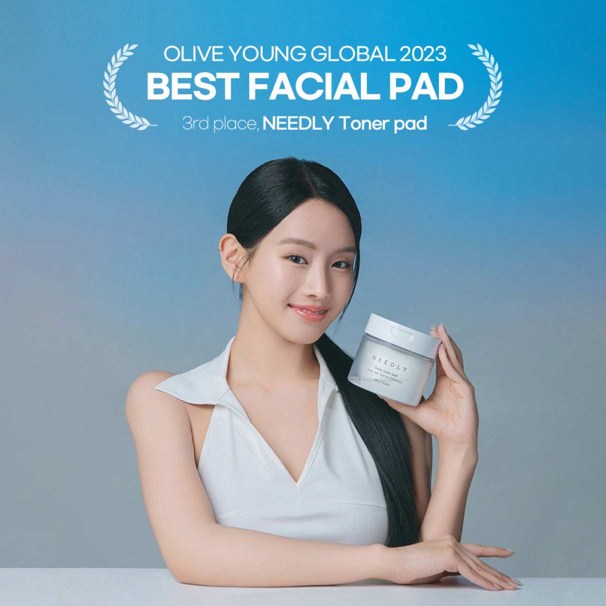 Giovane donna con lunghi capelli scuri e abito bianco sorride tenendo in mano un vasetto di NEEDLY Daily Toner Pad. Sullo sfondo sfumato blu e grigio, è visibile il testo: “Olive Young Global 2023 Best Facial Pad - 3rd place, NEEDLY Toner Pad”.