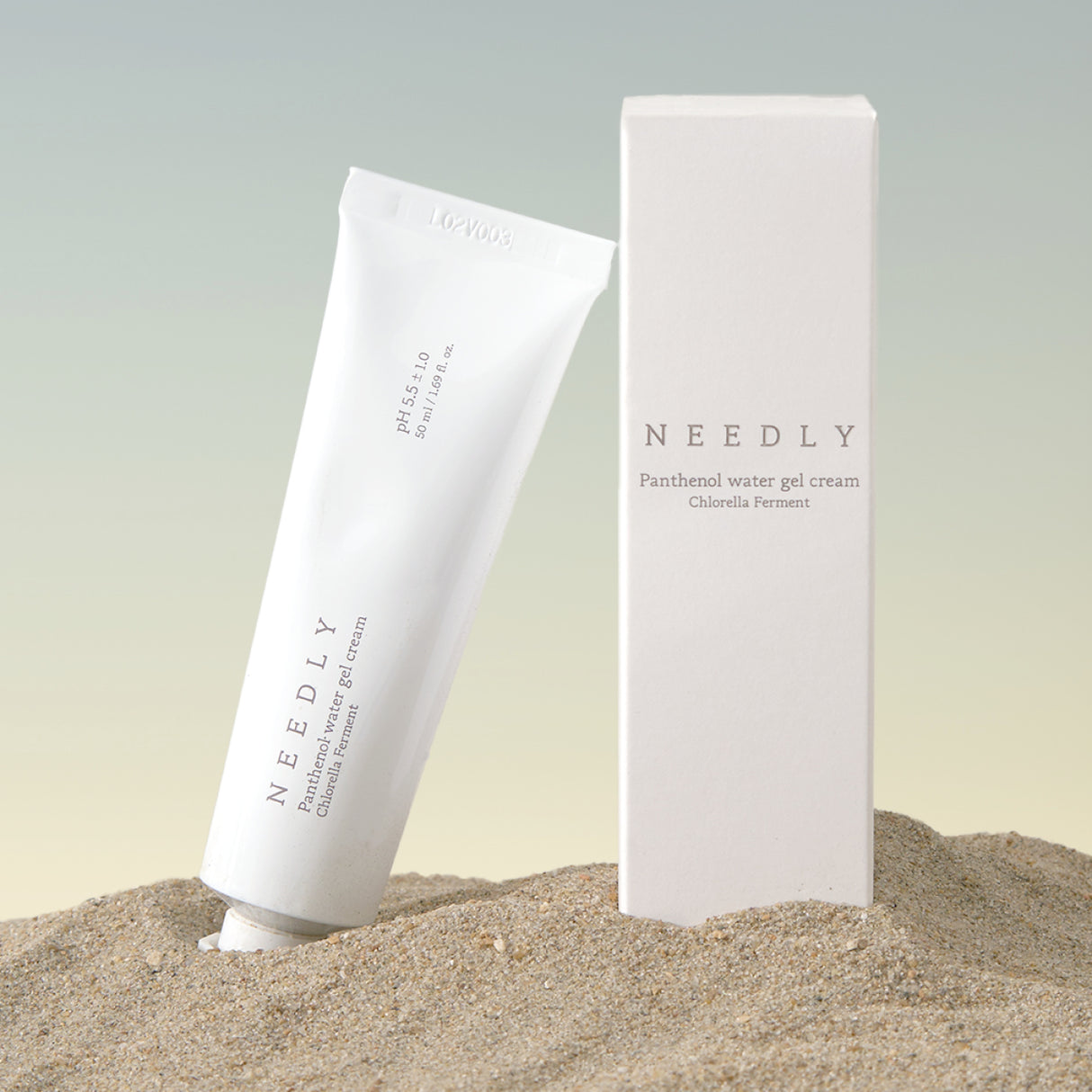 Il tubetto e la confezione della Needly Panthenol Water Gel Cream con Chlorella Ferment sono posizionati nella sabbia su sfondo sfumato beige-azzurro, creando un’atmosfera naturale e rilassante.