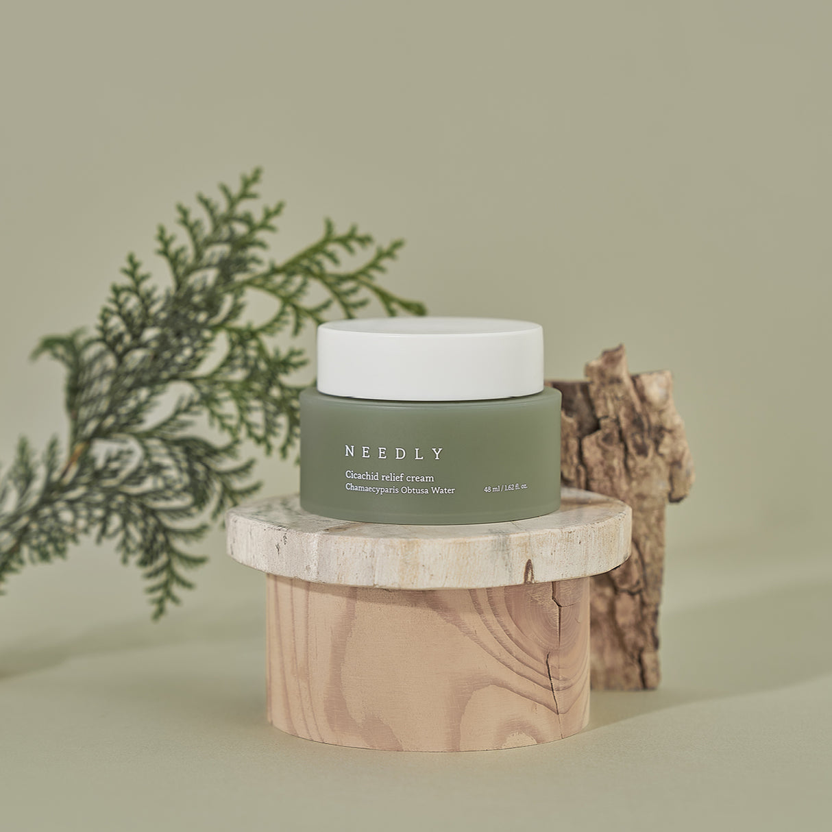 Crema Needly Cicachid Relief Cream da 48 ml con Chamaecyparis Obtusa Water, esposta su supporti in legno naturale con foglie e corteccia decorativa, ambientazione botanica su sfondo verde tenue.