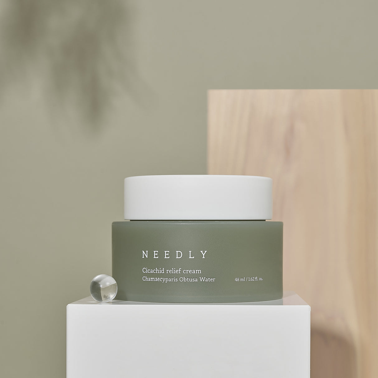 Crema viso Needly Cicachid Relief Cream 48 ml con Chamaecyparis Obtusa Water, packaging verde oliva con tappo bianco, esposta su piedistallo bianco con sfondo beige e decorazione in legno.