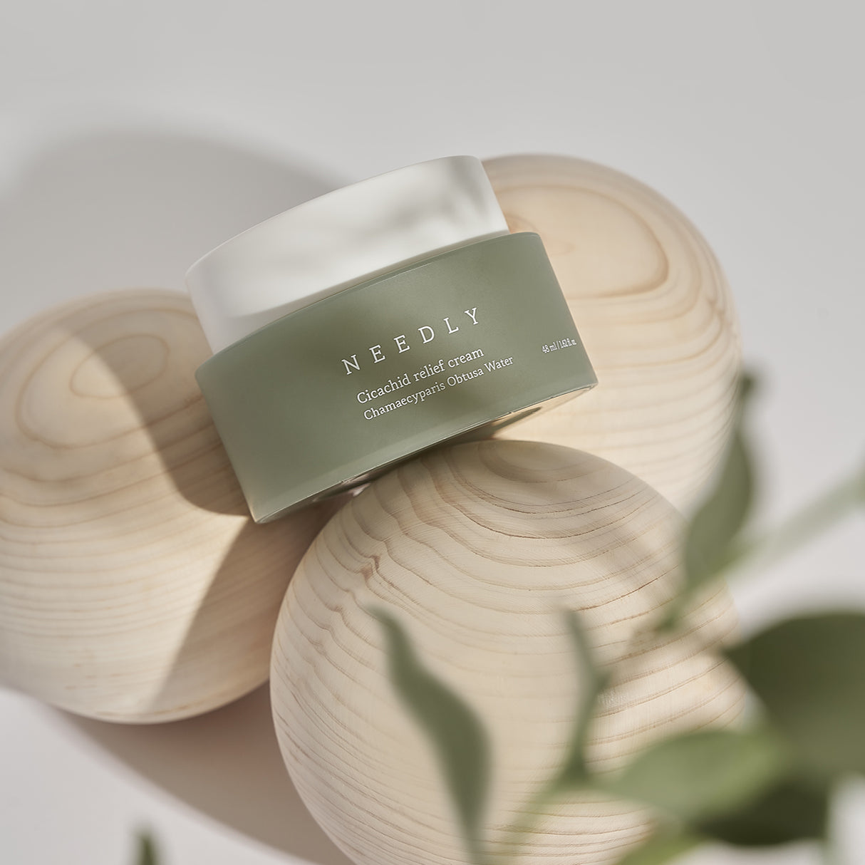 Crema viso Needly Cicachid Relief Cream 48 ml con Chamaecyparis Obtusa Water, confezione verde con tappo bianco, appoggiata su sfere di legno chiaro con decorazioni naturali in primo piano.