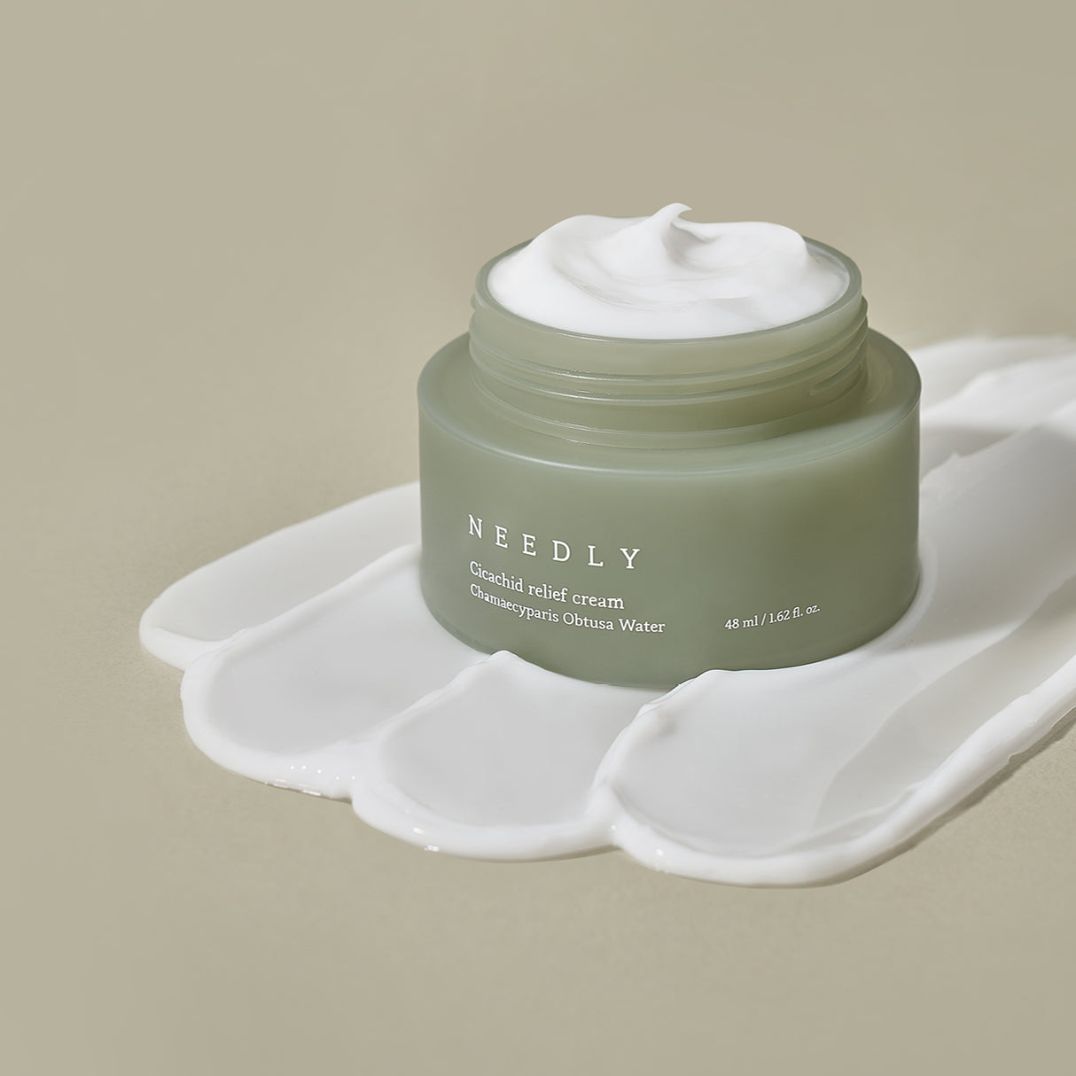 Crema viso Needly Cicachid Relief Cream 48 ml con Chamaecyparis Obtusa Water, vasetto verde su sfondo beige con texture del prodotto spalmata artisticamente alla base.