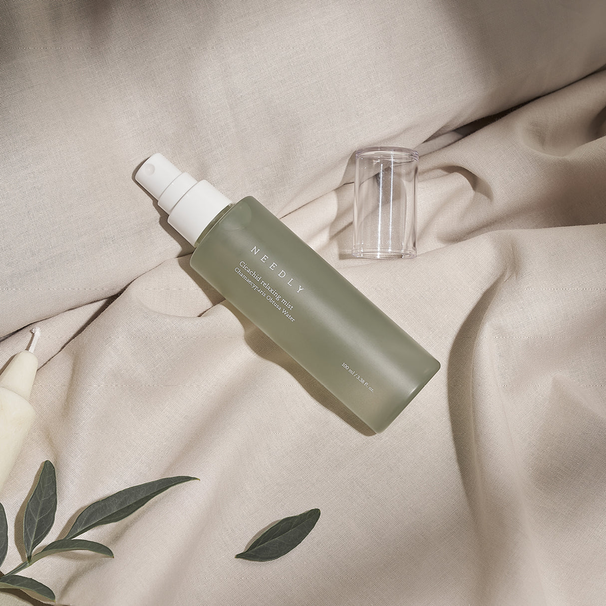 Flacone verde opaco della Needly Cicachid Relaxing Mist adagiato su un tessuto beige con tappo trasparente e decorazioni botaniche, illuminato da luce naturale.