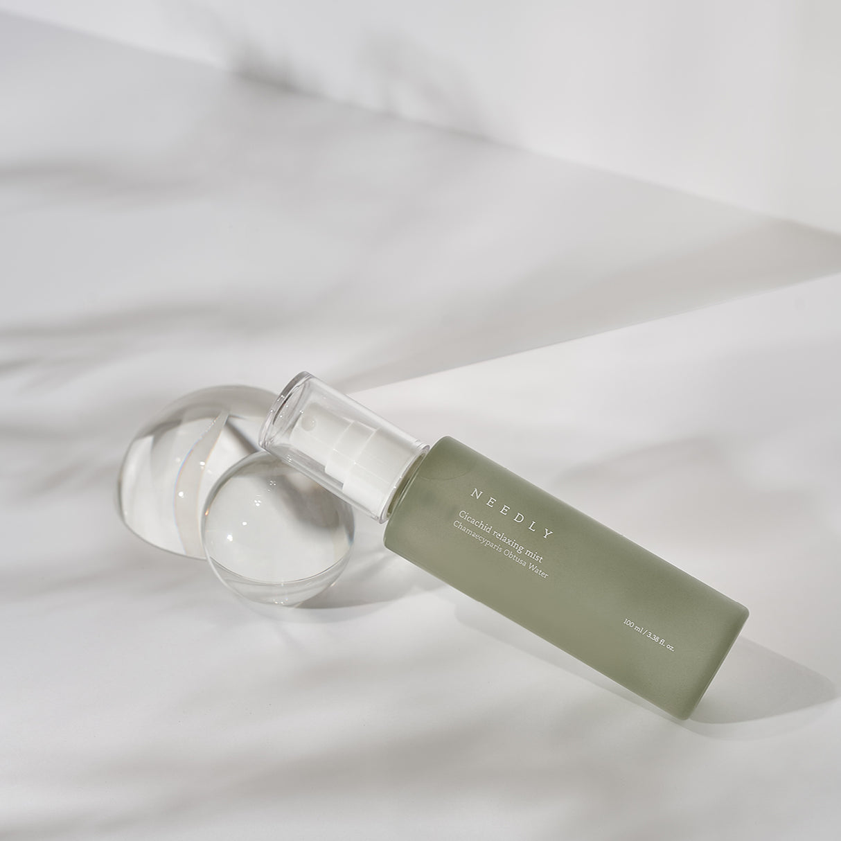 Flacone verde opaco della Needly Cicachid Relaxing Mist adagiato su una superficie bianca, accanto a due sfere di vetro trasparente. Lo sfondo è luminoso con giochi di ombre morbide.