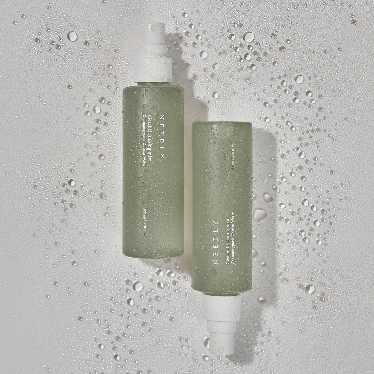 Due flaconi verdi opachi della Needly Cicachid Relaxing Mist adagiati su una superficie bianca bagnata, con gocce d’acqua che evocano freschezza e idratazione.