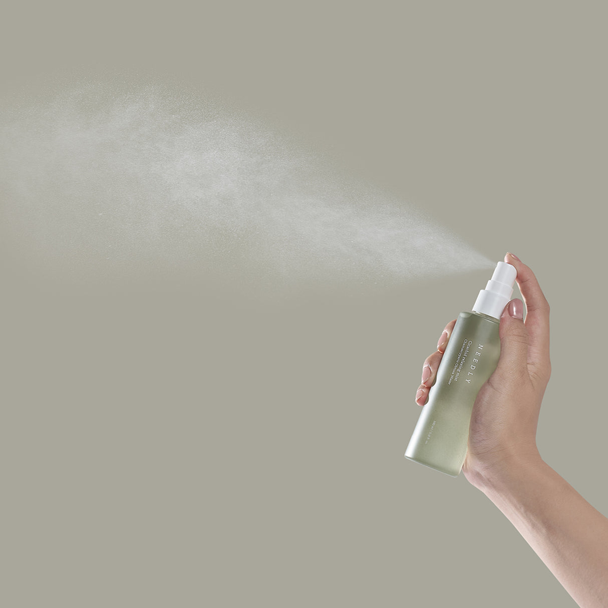 Mano che spruzza la Needly Cicachid Relaxing Mist, mostrando una nuvola sottile e diffusa di prodotto su sfondo beige, enfatizzando l'effetto rinfrescante e leggero dello spray viso.