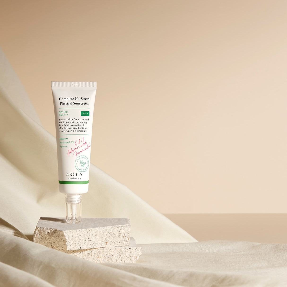 Tubetto di crema solare AXIS-Y Complete No-Stress in posizione verticale, appoggiato su blocchi di pietra beige con sfondo in tessuto chiaro e luce naturale.