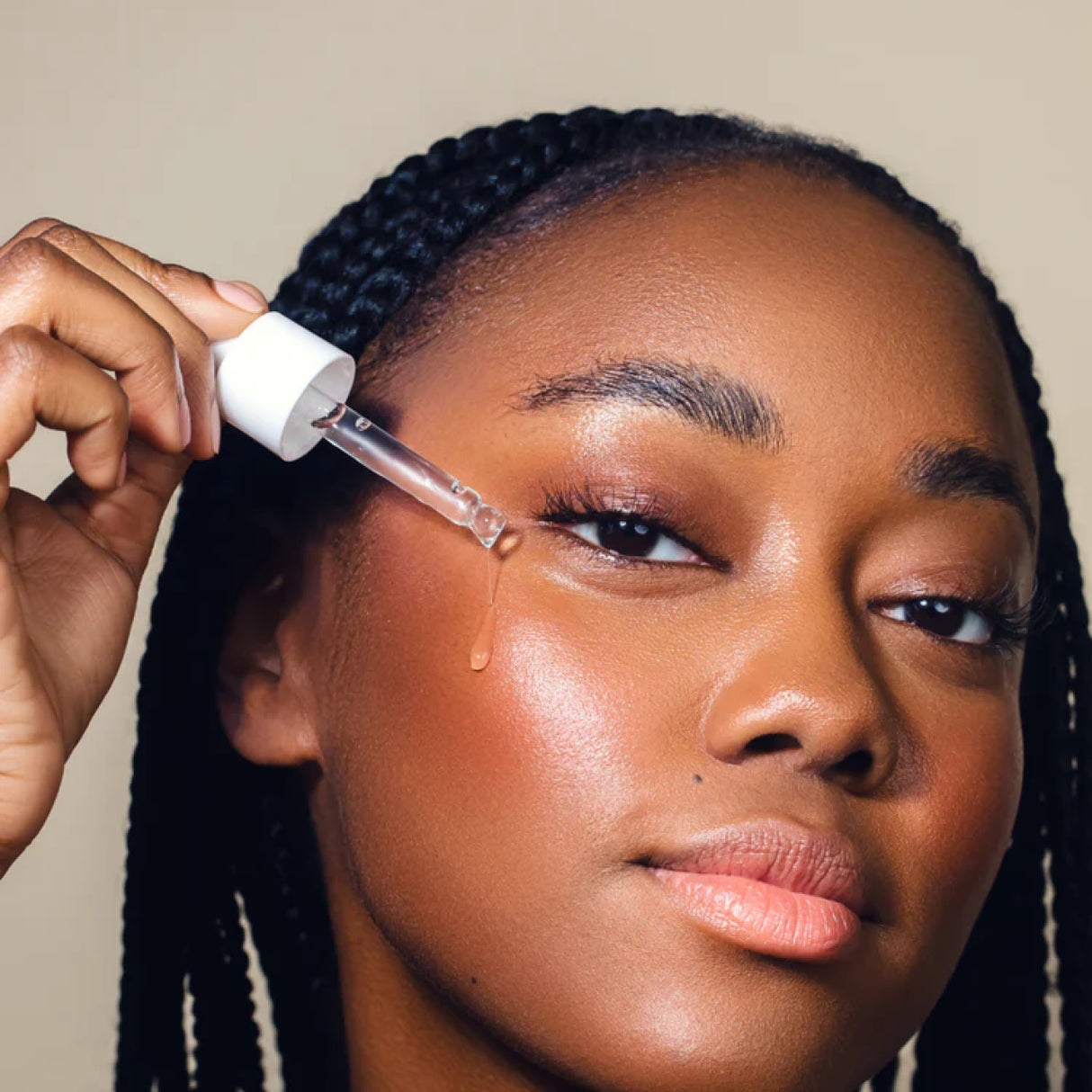 Primo piano di una persona che applica l’essenza viso con una pipetta, lasciando cadere una goccia di prodotto sulla guancia, su sfondo neutro.