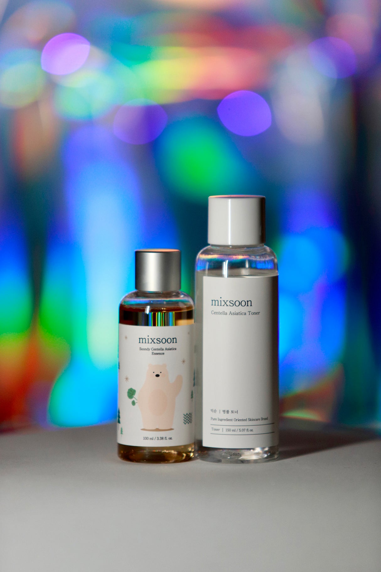 Due flaconi trasparenti del brand Mixsoon su sfondo sfocato dai colori vivaci: a sinistra il Beauty Centella Asiatica Essence con illustrazione di un orsetto, a destra il Centella Asiatica Toner con etichetta minimalista bianca.