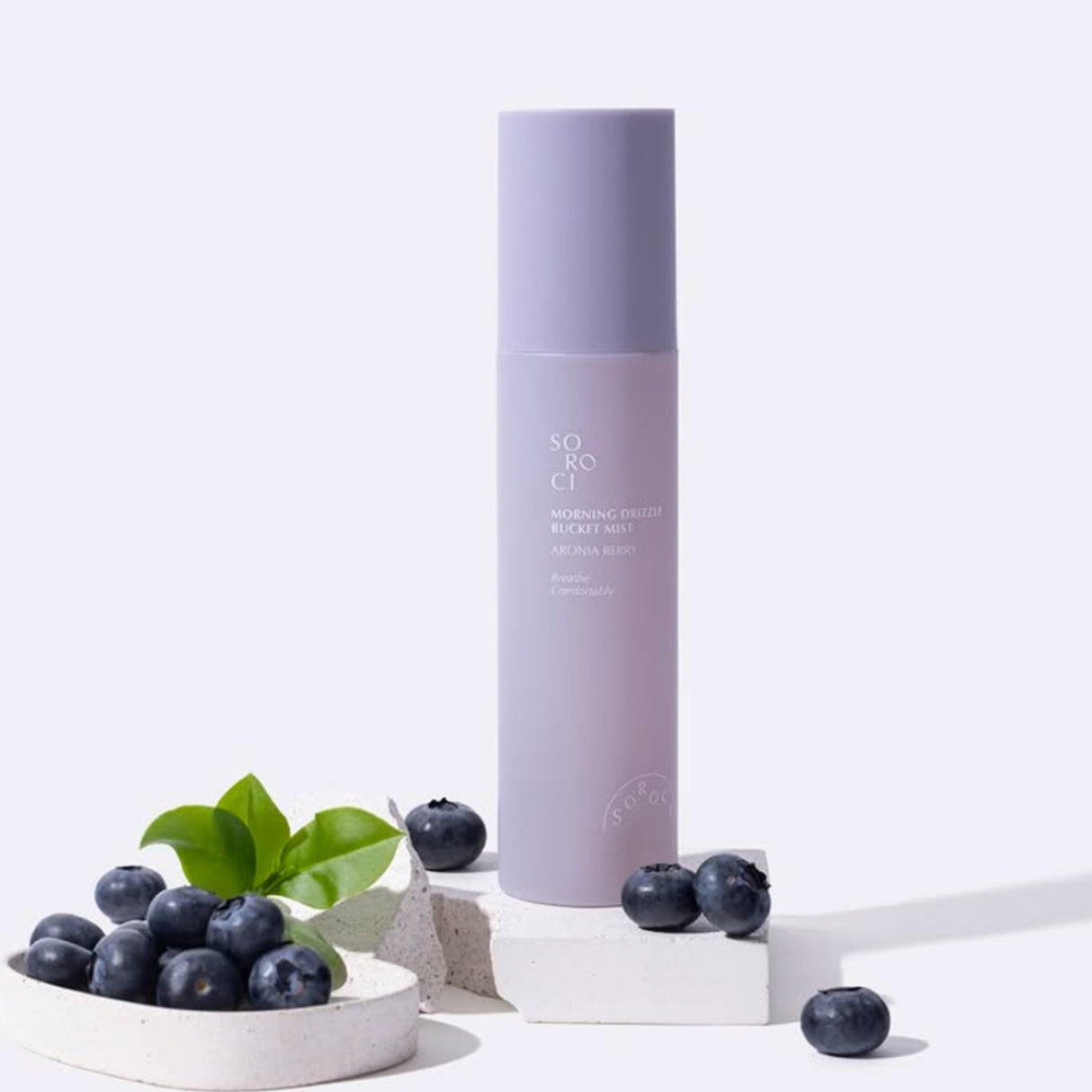 Flacone verticale del mist viso SOROCI Morning Drizzle Bucket Mist alla aronia berry, posizionato su blocchi chiari con mirtilli freschi e foglie verdi, su sfondo bianco.