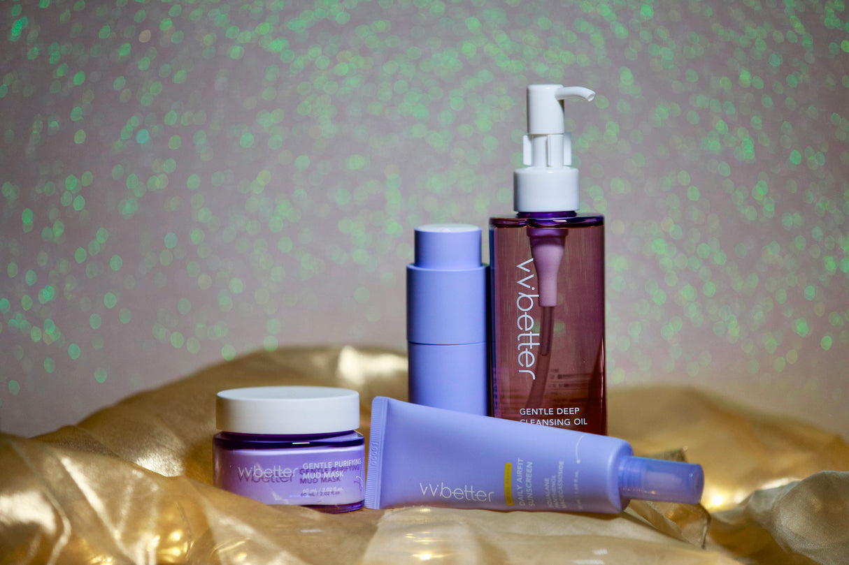 Set skincare VVBetter con Gentle Deep Cleansing Oil, Gentle Purifying Mud Mask, Daily Airfit Sunscreen e altri trattamenti viso in packaging lilla e viola, esposti su tessuto dorato con sfondo glitterato verde perlescente.
