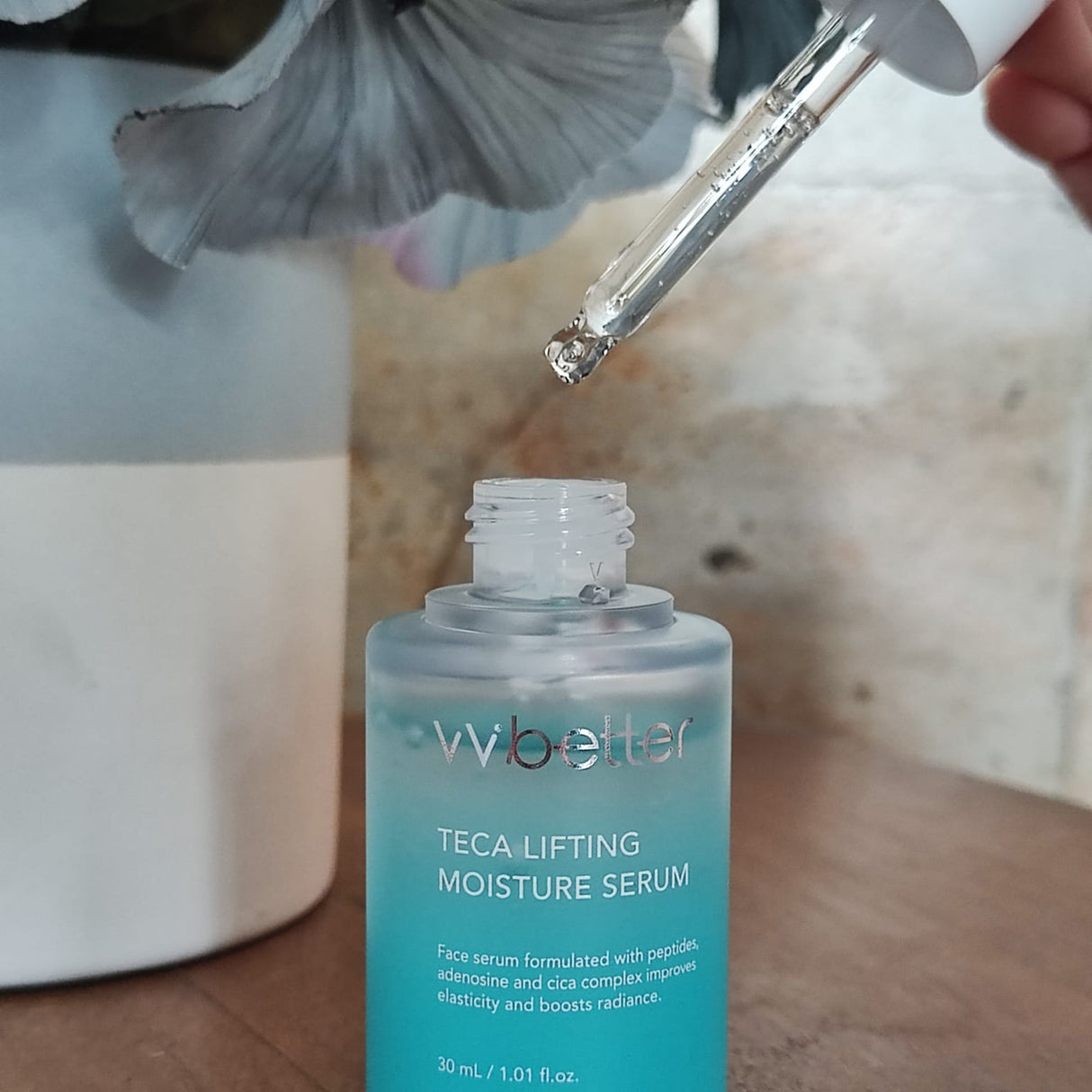 Contagocce che applica il siero Wbetter TECA Lifting Moisture in un flacone aperto da 30 ml, su sfondo naturale con fiore decorativo
