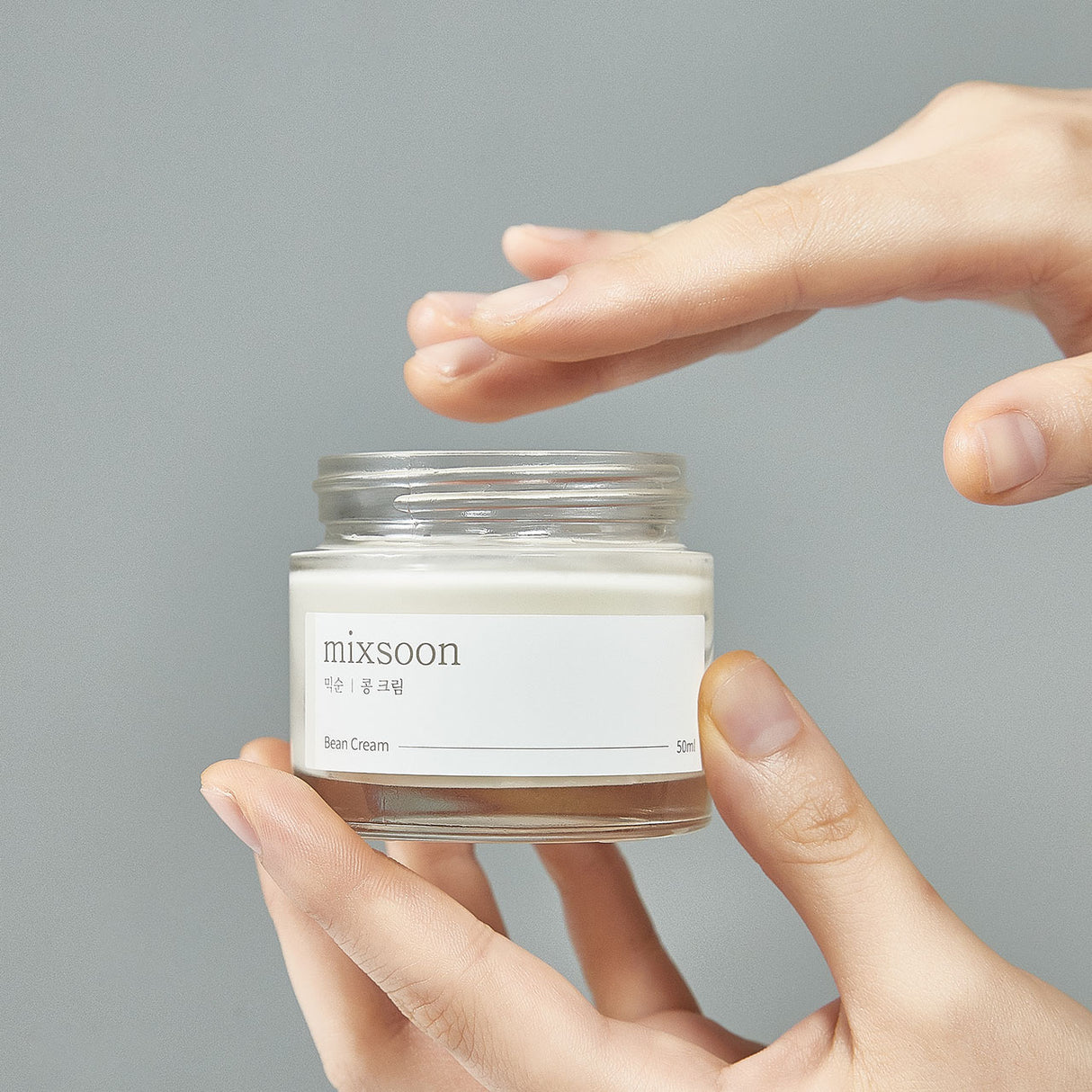 "Mani che tengono e prelevano delicatamente la crema da un barattolo aperto di Mixsoon Bean Cream con sfondo grigio chiaro."