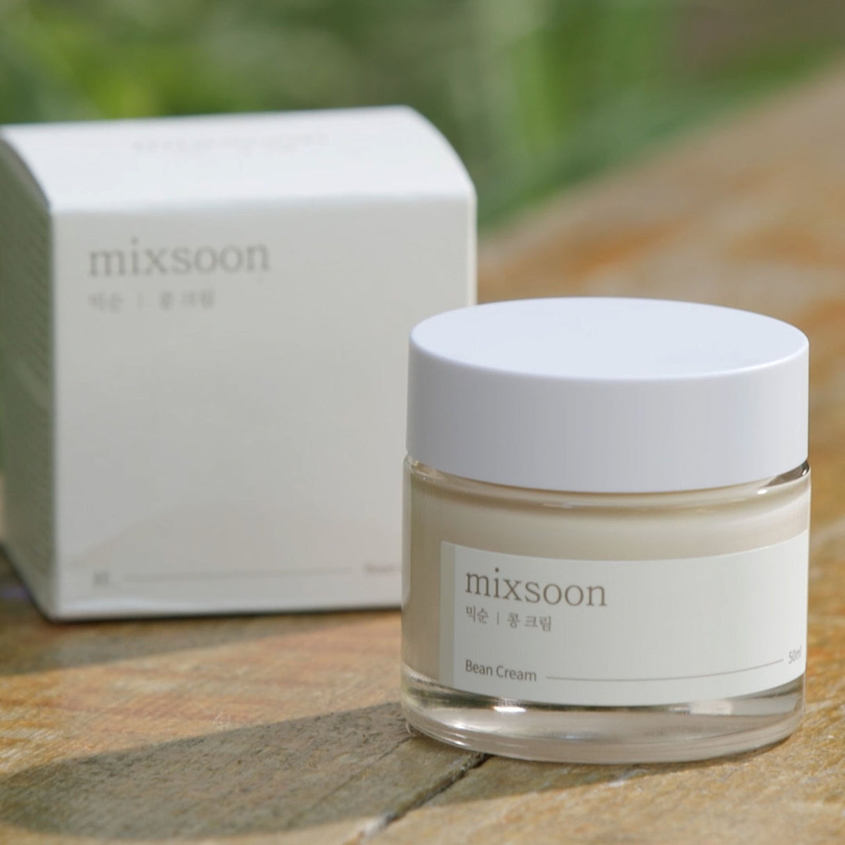 Barattolo di Mixsoon Bean Cream da 50 ml con tappo bianco, posizionato su una superficie in legno accanto alla confezione originale, con sfondo sfocato di elementi naturali."