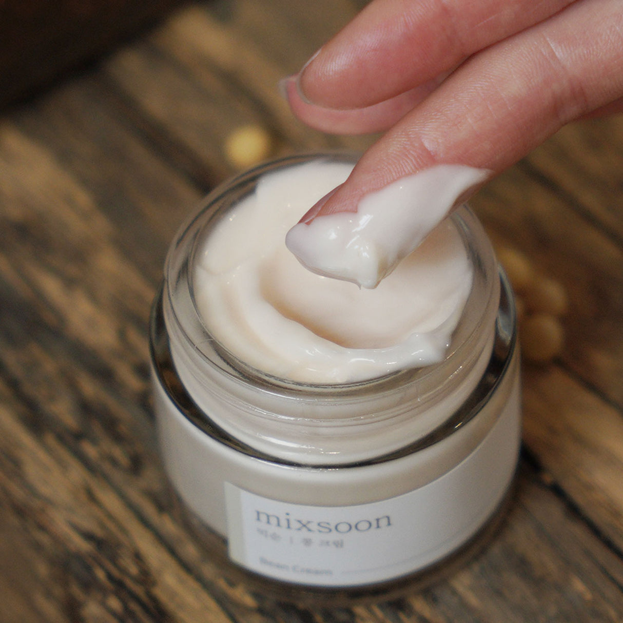 "Primo piano di una mano che preleva una dose di Mixsoon Bean Cream da un barattolo aperto, mostrando la consistenza ricca e cremosa del prodotto su sfondo in legno rustico.