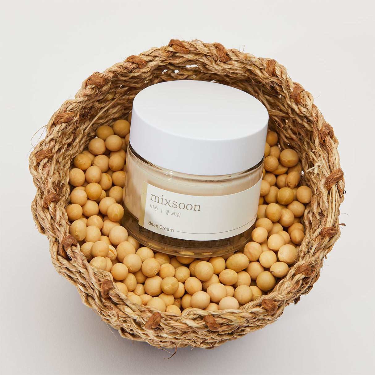 Vasetto di Mixsoon Bean Cream posizionato all’interno di un cestino intrecciato, circondato da fagioli di soia secchi su sfondo bianco.