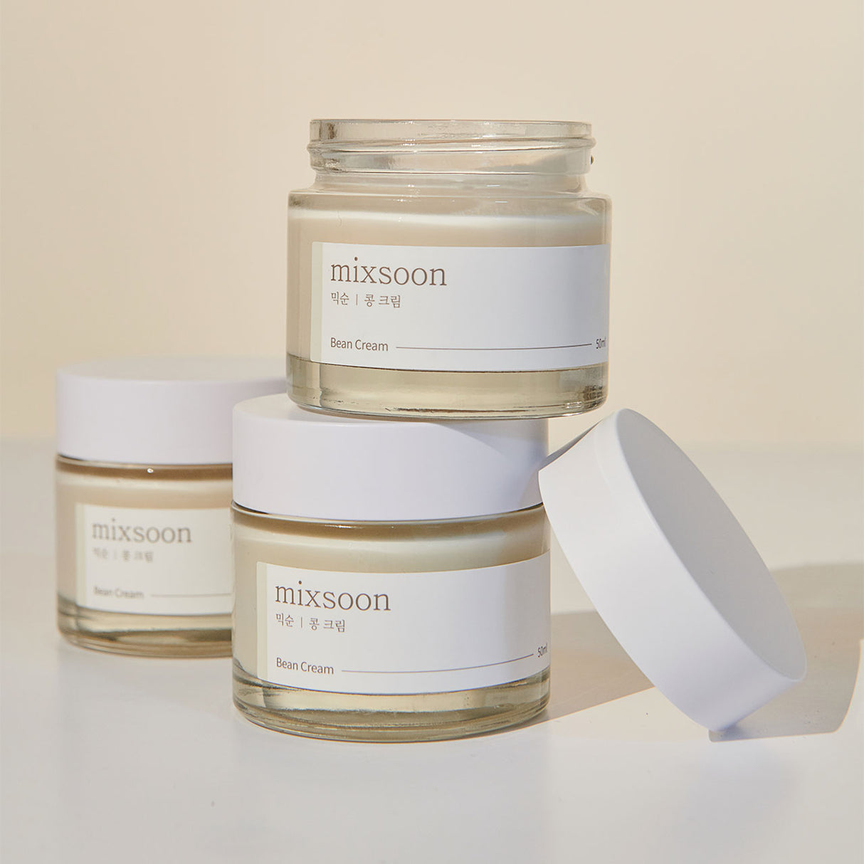 Tre vasetti di Mixsoon Bean Cream disposti su una superficie chiara, con uno impilato sopra un altro e un coperchio appoggiato lateralmente, illuminati da una luce naturale calda."