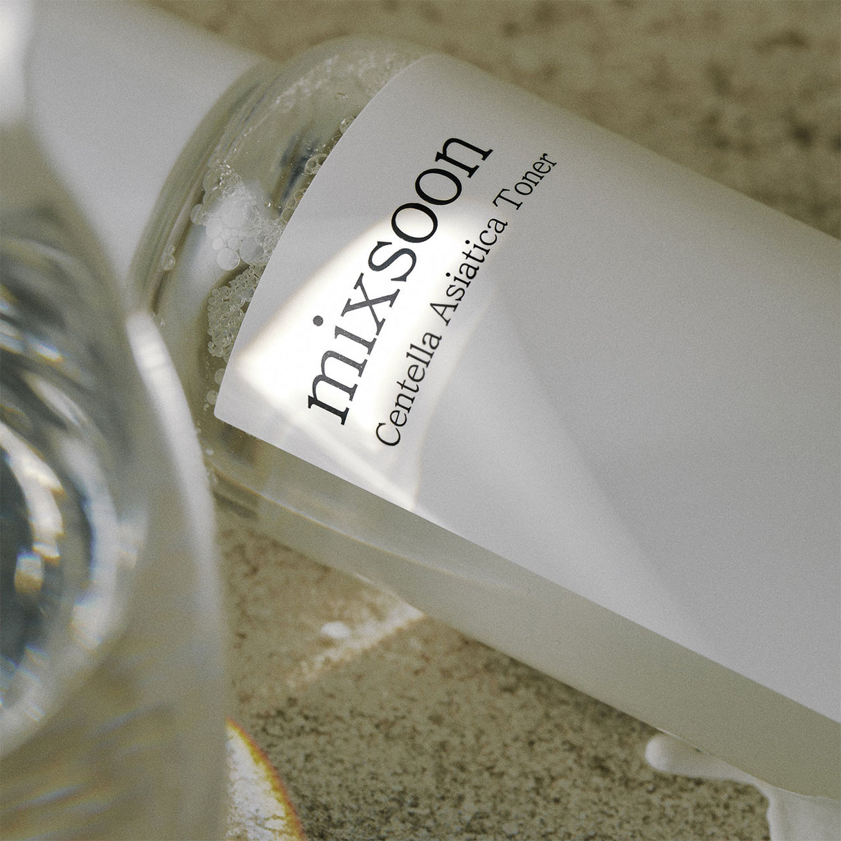 Primo piano della bottiglia Mixsoon Centella Asiatica Toner adagiata su una superficie naturale. La luce morbida e i riflessi nell’acqua enfatizzano la purezza e la trasparenza del tonico calmante per la pelle.