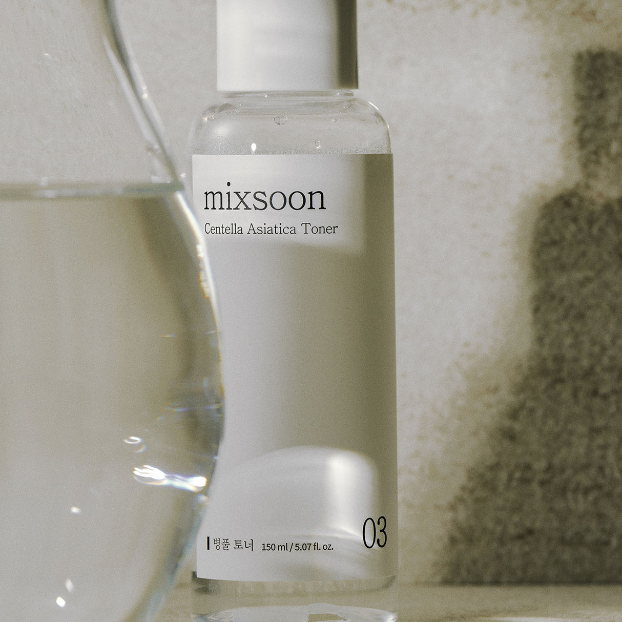 Flacone minimalista da 150 ml di Mixsoon Centella Asiatica Toner posizionato su una superficie in pietra, con luce naturale e un bicchiere d'acqua sfocato in primo piano che richiama purezza e idratazione.