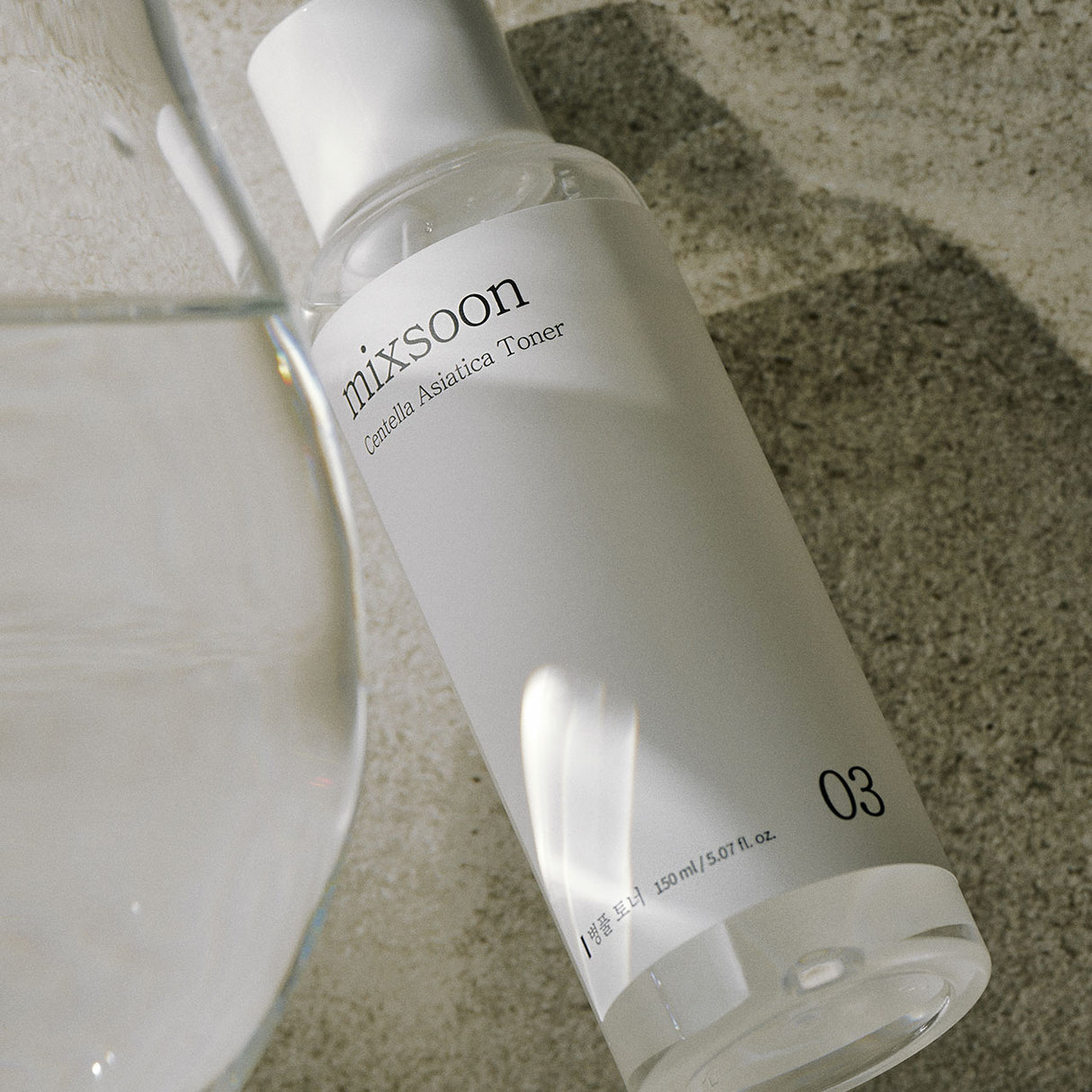 Flacone da 150 ml di Mixsoon Centella Asiatica Toner disteso su una superficie in pietra grezza, affiancato da un bicchiere d’acqua sfocato. L’inquadratura obliqua e la luce naturale trasmettono freschezza e semplicità.