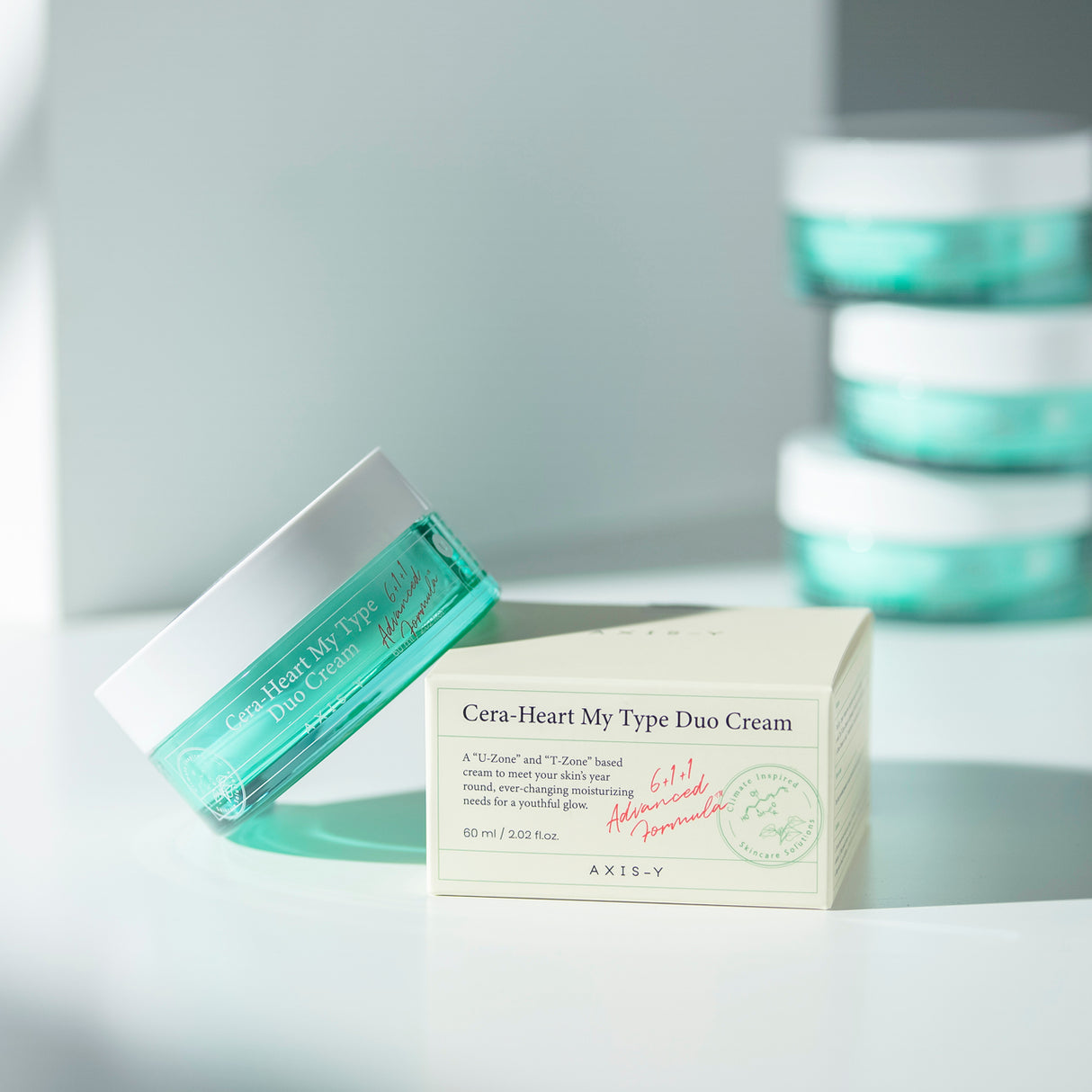 Vasetto inclinato di crema viso AXIS-Y Cera-Heart My Type Duo Cream accanto alla confezione in cartoncino, su sfondo chiaro con altri vasetti sfocati sullo sfondo.