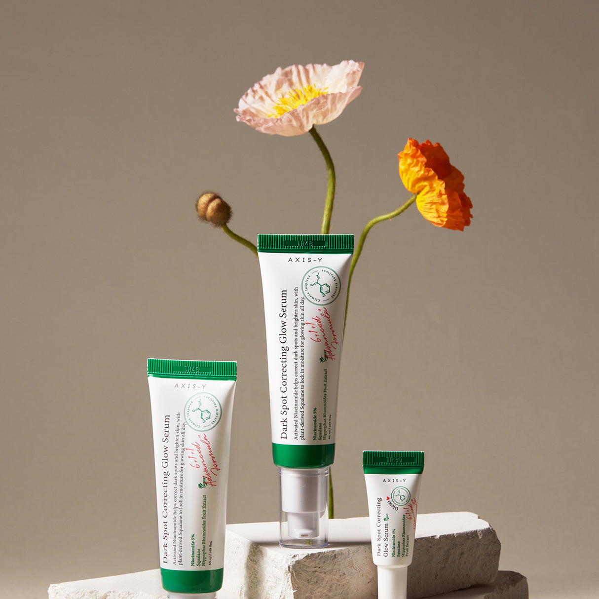 Tre formati del siero AXIS-Y Dark Spot Correcting Glow Serum disposti su blocchi bianchi in pietra con due fiori colorati sullo sfondo beige, creando una composizione elegante e naturale.