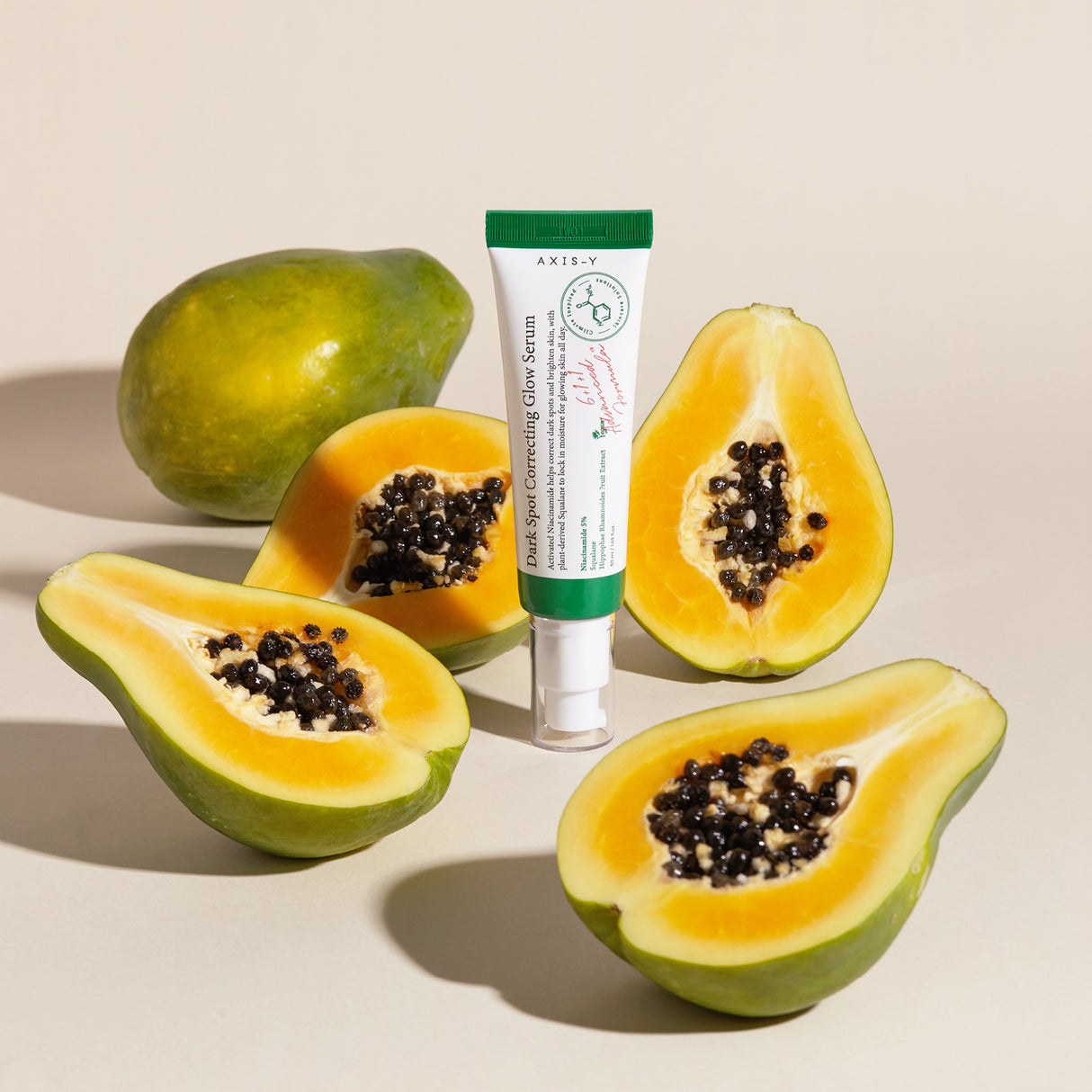 Tubo di siero AXIS-Y Dark Spot Correcting Glow Serum in piedi su sfondo chiaro, circondato da papaye fresche intere e tagliate a metà, con semi neri in evidenza.