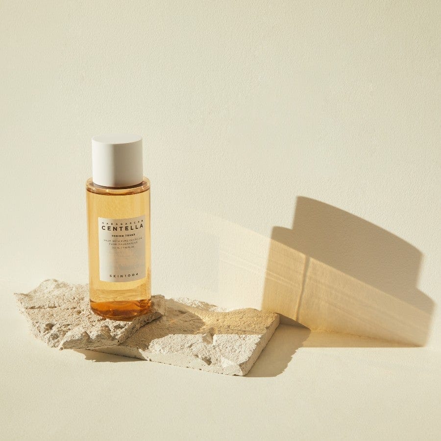 Toner viso alla centella asiatica FOODAHOLIC su fondo neutro con base in pietra, flacone in vetro trasparente con tappo bianco, skincare coreana idrata