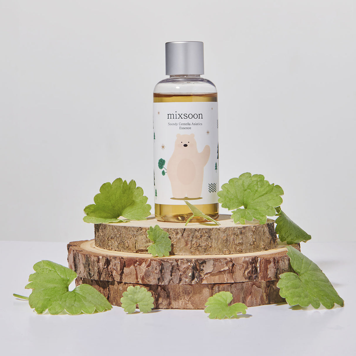 Flacone di Mixsoon Soondy Centella Asiatica Essence posizionato su tre dischi di legno grezzo impilati, circondato da foglie fresche di centella asiatica. Lo sfondo è bianco neutro, che mette in risalto gli elementi naturali del prodotto.