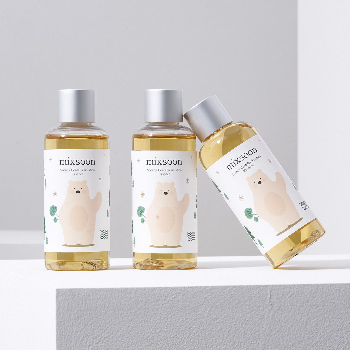 Tre flaconi di Mixsoon Soondy Centella Asiatica Essence con etichetta decorata da un orsetto beige stilizzato. Due flaconi sono in piedi e uno è inclinato appoggiandosi lateralmente. Sfondo chiaro e illuminazione soffusa per un effetto pulito ed elegante.