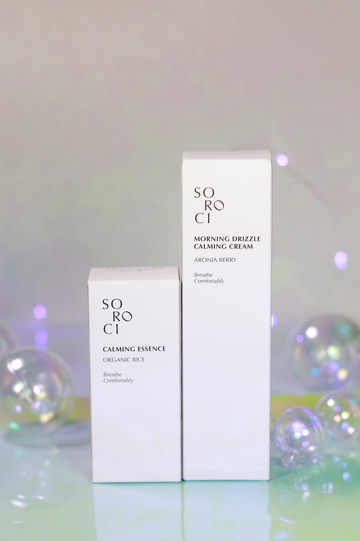 Due confezioni bianche dei prodotti Soroci su sfondo color pastello con bolle trasparenti: a sinistra "Calming Essence Organic Rice", a destra "Morning Drizzle Calming Cream Aronia Berry", disposte in verticale.