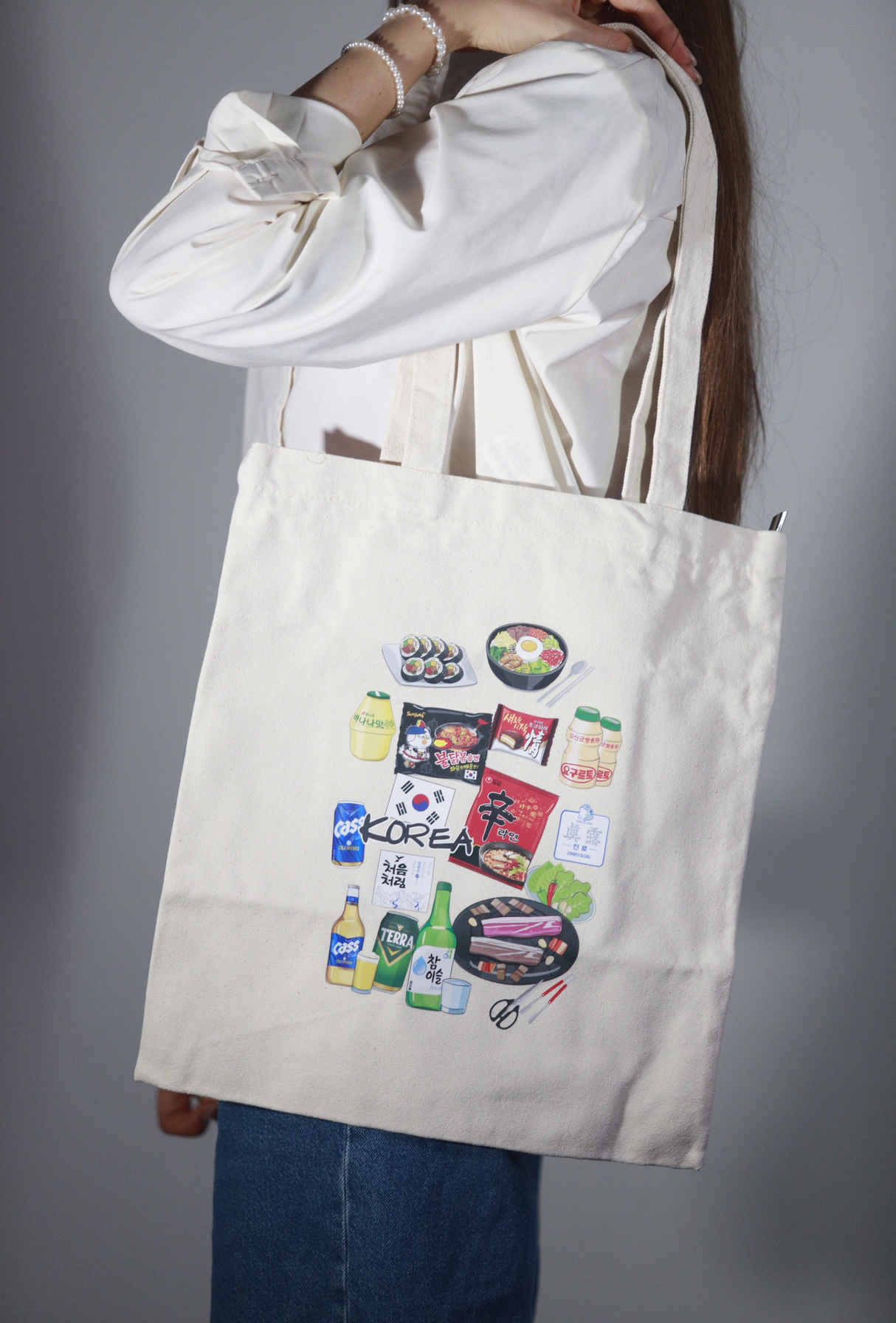 Shopper bag in tessuto color panna portata a spalla da una persona, con grafiche illustrate di ramen, sushi, bevande e snack coreani su sfondo neutro