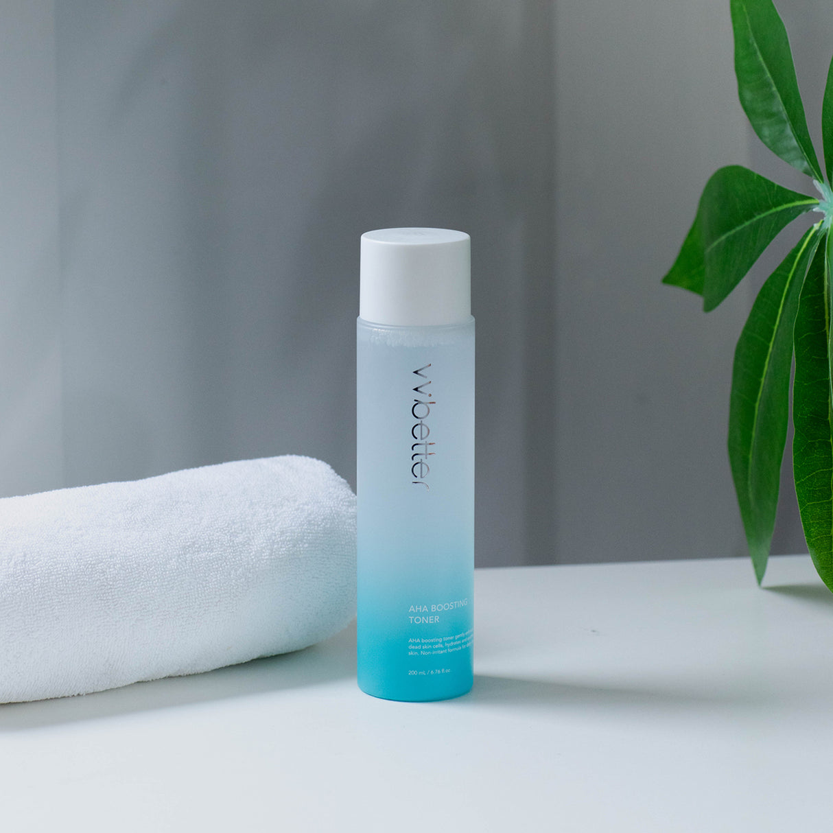 Flacone sfumato azzurro da 200 ml di AHA Boosting Toner del marchio vvbetter, con tappo bianco, posizionato su una superficie bianca accanto a un asciugamano arrotolato e una pianta verde, su sfondo neutro.