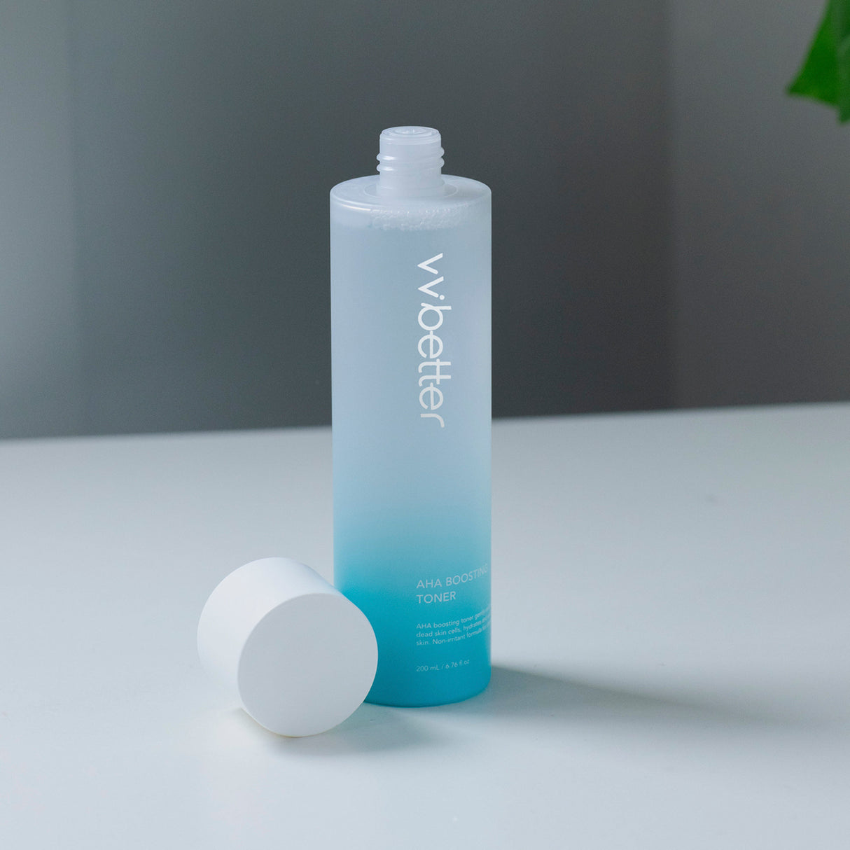 Flacone sfumato azzurro del tonico "AHA Boosting Toner" di vvbetter da 200 ml, con tappo bianco rimosso appoggiato accanto, su sfondo minimal bianco e grigio.