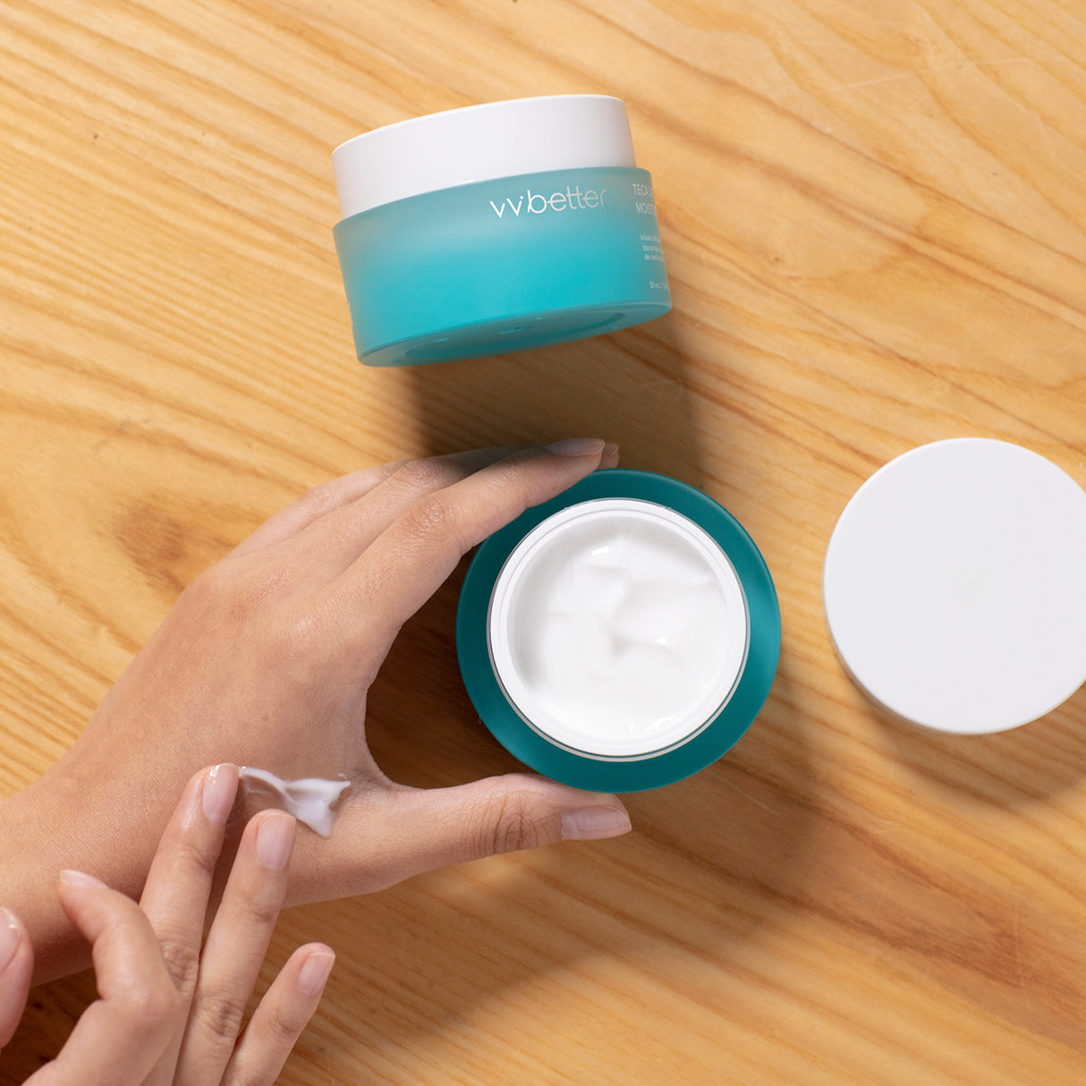Una mano preleva e applica la vvbetter Teca Lifting Moisture Cream da un vasetto aperto color azzurro su un piano in legno chiaro. La consistenza cremosa bianca della crema è ben visibile, trasmettendo una sensazione di idratazione e cura della pelle.