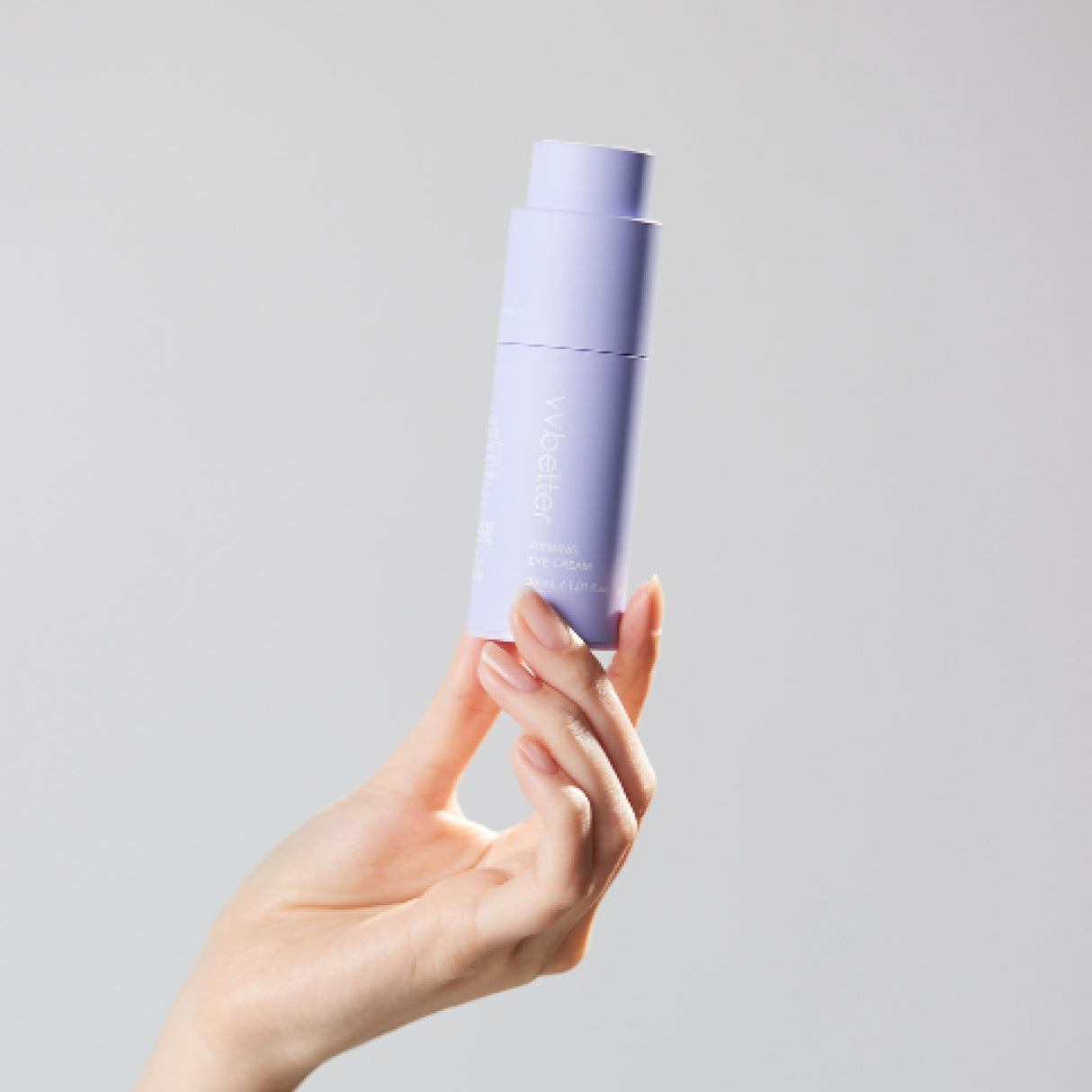 Una mano solleva un flacone color lavanda del vvbetter Firming Eye Cream da 30 ml / 1.01 fl.oz., su sfondo neutro chiaro.